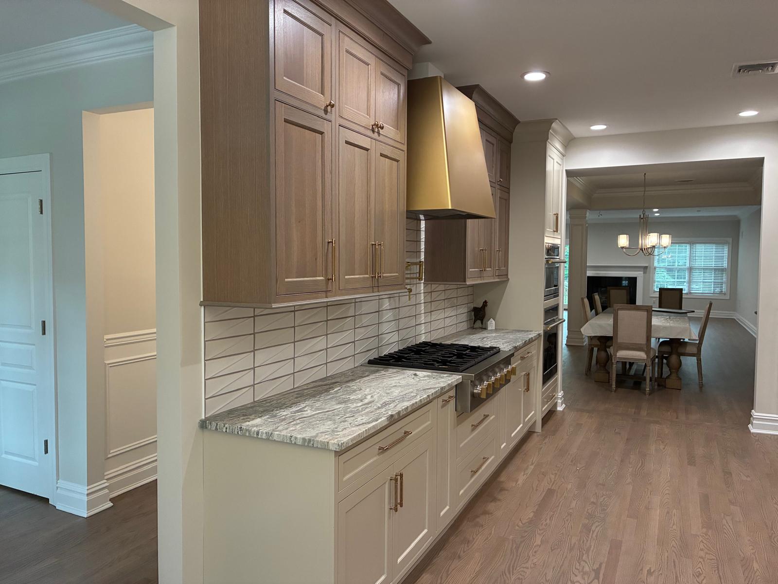 Completed custom kitchen with wood cabinetry, brass range hood, stone counters, and open dining area